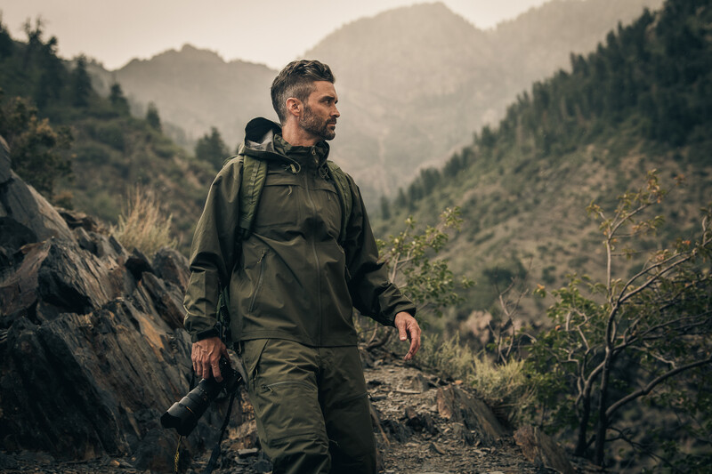 Hiker in olive jacket walking on mountain trail surrounded by forested slopes.