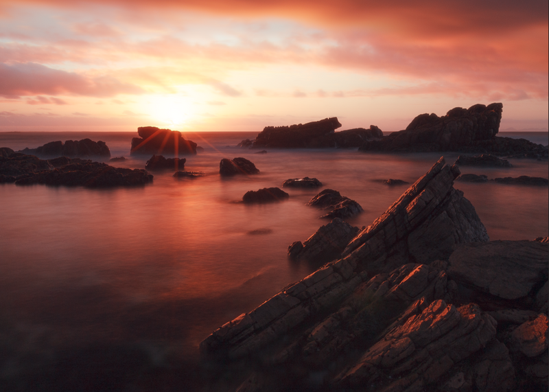 Rocky coastal landscape at sunset with warm orange sky reflecting on calm water and silhouetted rock formations.