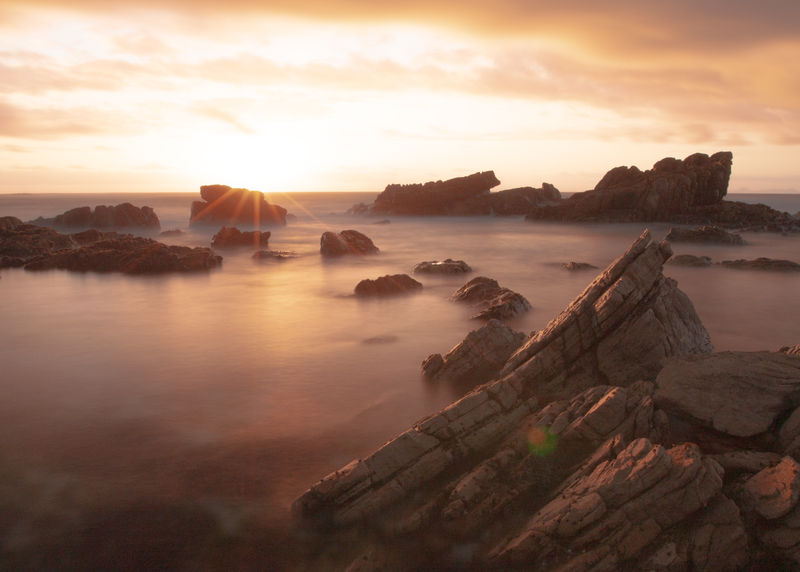 Coastal landscape at sunrise with silhouetted rock formations and driftwood in calm, misty water.