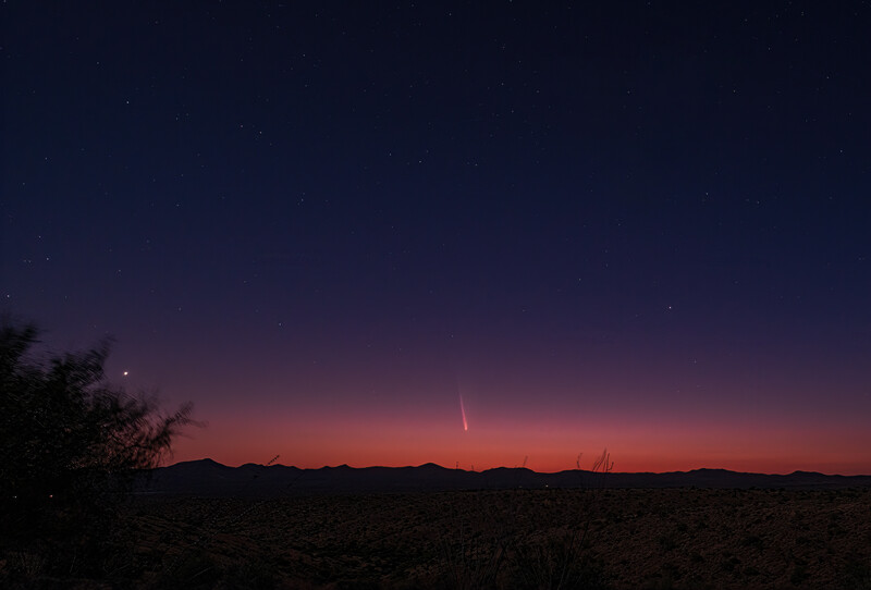 Desert landscape at twilight with silhouetted mountains and a bright meteor streaking across the darkening sky.