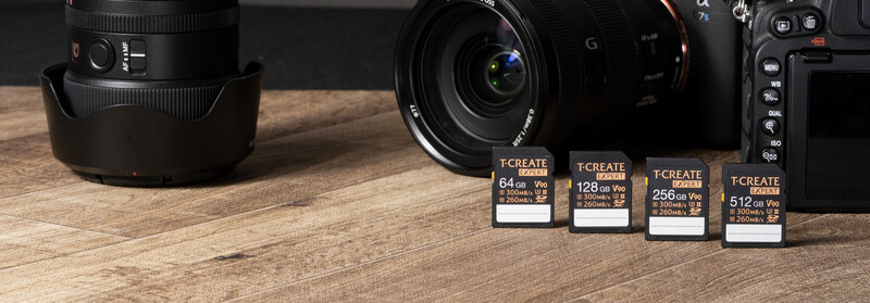 Four T.CREATE SD memory cards displayed with camera lenses and a mirrorless camera body on wooden surface.