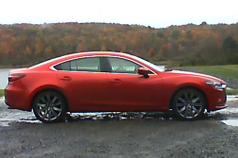 Red sedan photographed in profile on a wet asphalt surface with autumn foliage in the background.