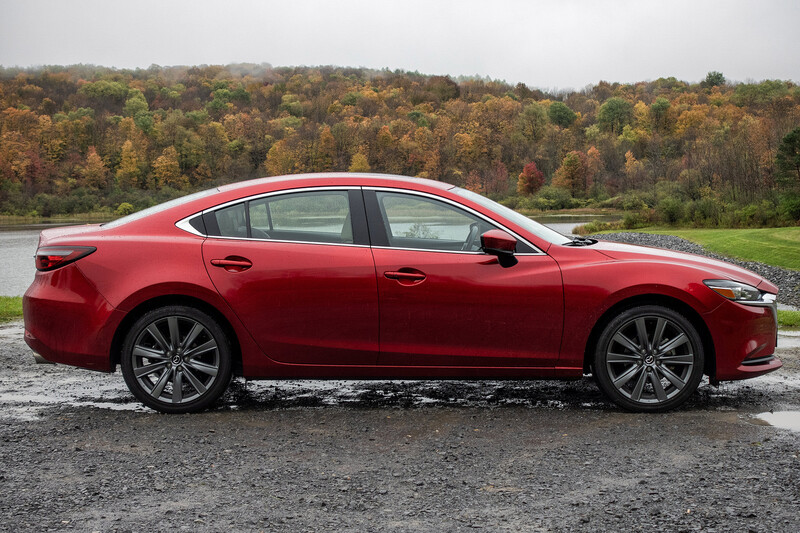 Red sedan parked on wet asphalt with autumn forest and water in background.