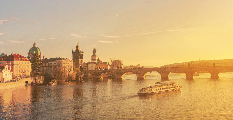Historic European riverside city at sunset with stone bridge and church spires reflected in calm water.