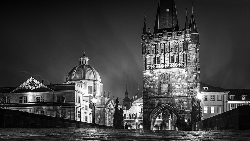 Gothic tower with ornate stonework illuminated at night beside a domed building across a river.