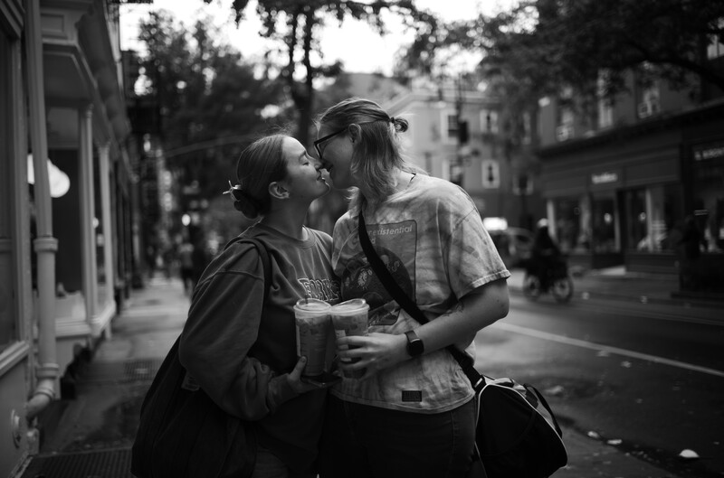 Two people sharing an intimate moment on a city street, one wearing a cap and holding a beverage.
