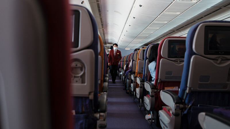 Interior of an aircraft cabin with rows of seats extending down the aisle, a flight attendant in red uniform visible in the distance.
