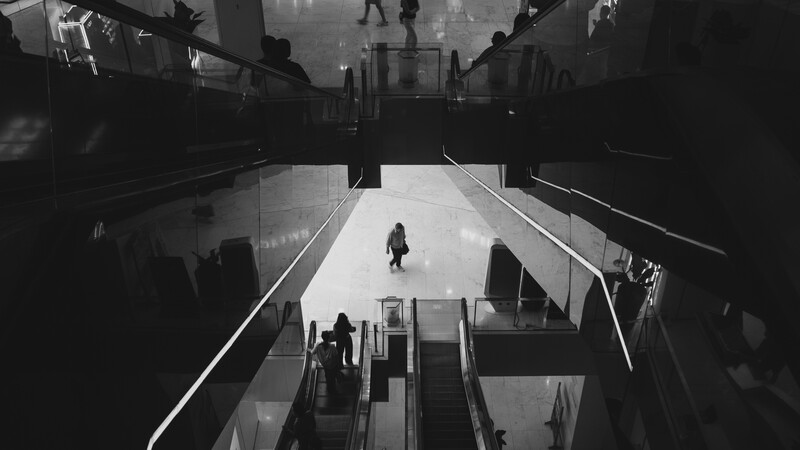 Overhead view of a multi-level subway or train station with a lone figure standing on the central platform surrounded by escalators and architectural geometry.