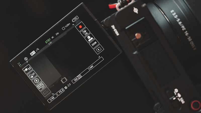 Camera monitor displaying menu interface with lens attached, photographed against dark background.