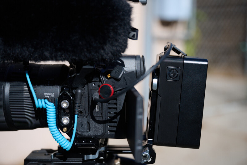 Professional cinema camera with attached microphone, monitor, and blue audio cable in outdoor setting.