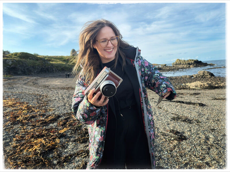 Photographer holding vintage film camera on a rocky beach with coastal landscape behind her.