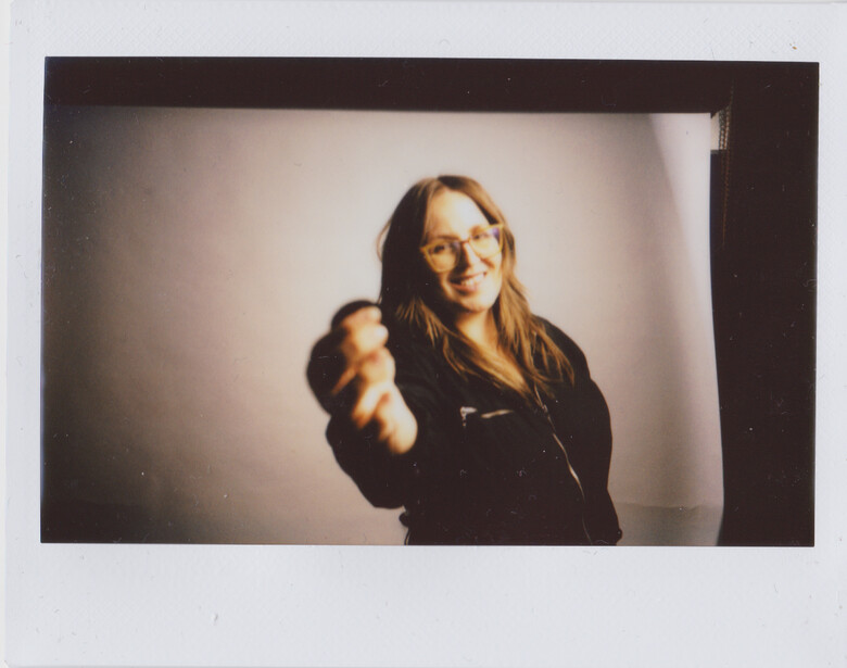 Person in dark jacket pointing toward camera with warm smile against neutral backdrop.
