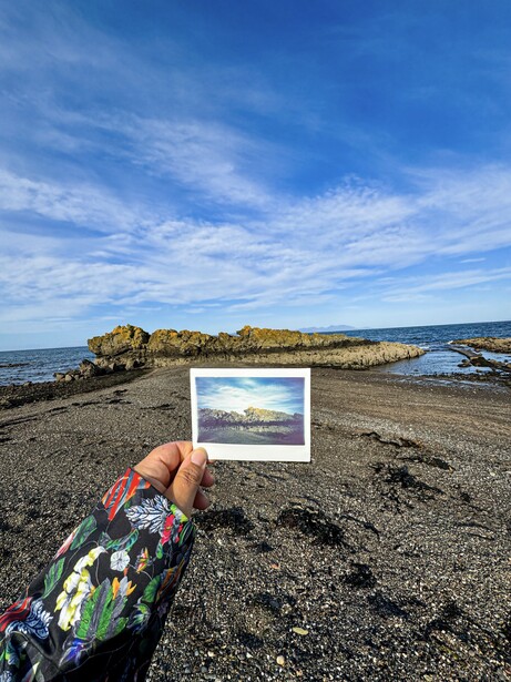 Hand holding a landscape photograph while standing on a rocky beach with matching scenery in background.