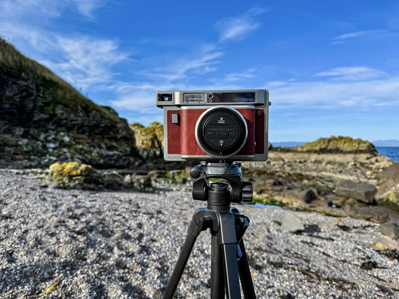 Red rangefinder camera mounted on tripod overlooking rocky coastal landscape with blue sky.