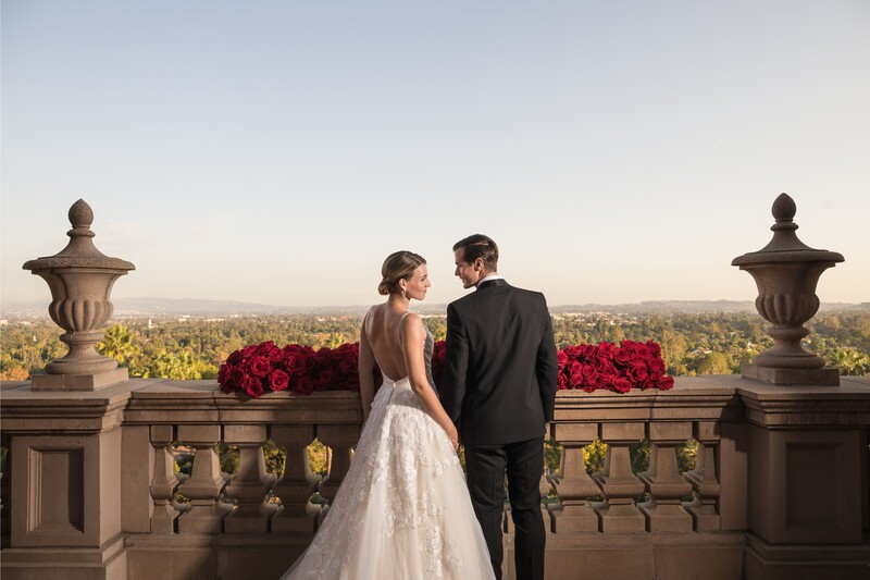 Bride and groom stand at a stone balustrade overlooking a sprawling city landscape at golden hour.