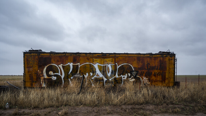 Rusted railroad boxcar with large white graffiti tag sits abandoned in sparse grassland under overcast sky.