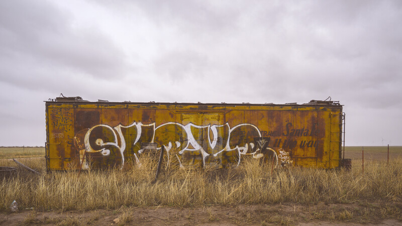 Abandoned railway boxcar with white graffiti in an arid grassland under overcast sky.