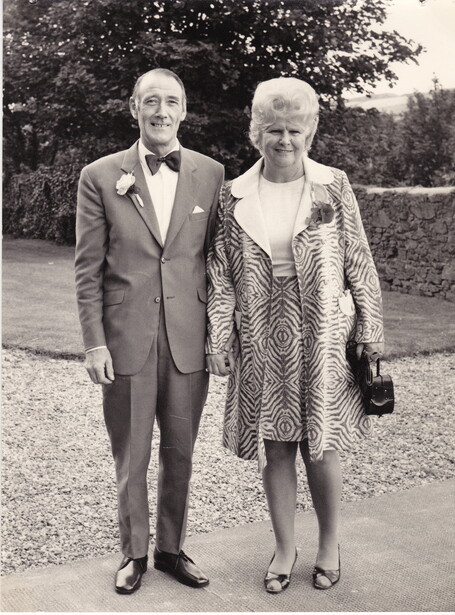 Man in suit and woman in patterned dress walk together on a gravel path lined with hedges.