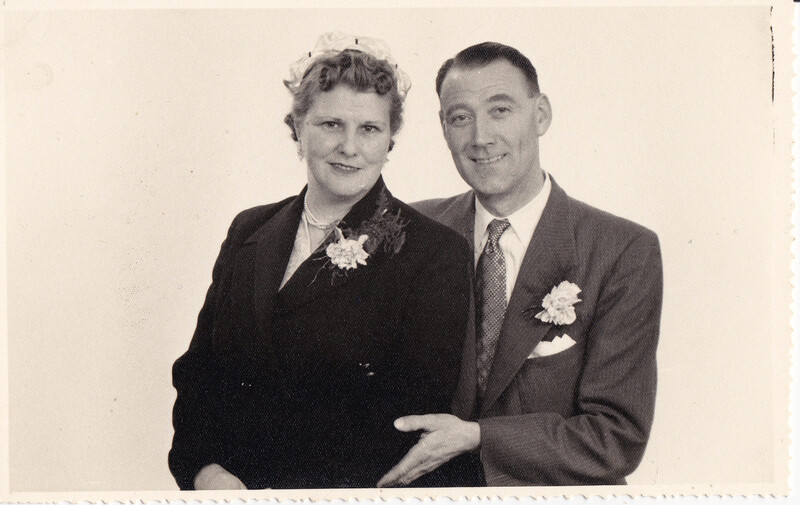 Formal portrait of a man and woman wearing corsages, posed against a neutral background.