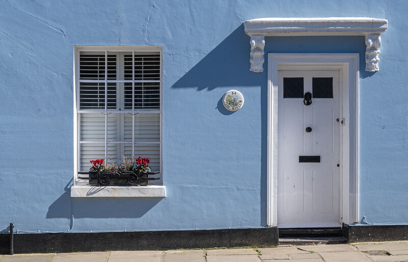 Minimalist facade of a bright blue house with white door, shuttered window, and red flowers.