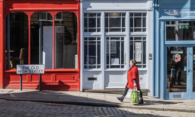 Person in red coat walking past row of colorful storefronts on historic street.