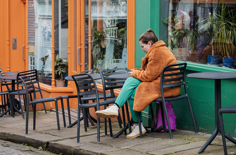 Person in tan cardigan sitting at outdoor café table with black metal chairs and colorful storefront behind.