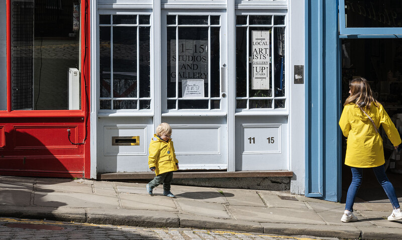 Two children in yellow jackets walking on a sidewalk in front of a storefront with colorful painted frames.