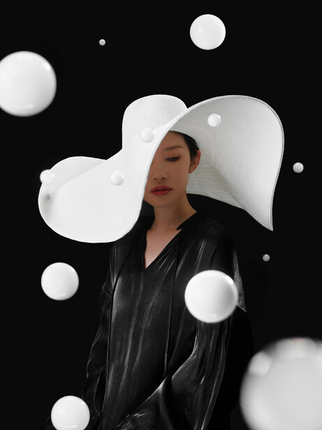 Woman in black jacket wearing oversized white sculptural hat against black background with white spheres.