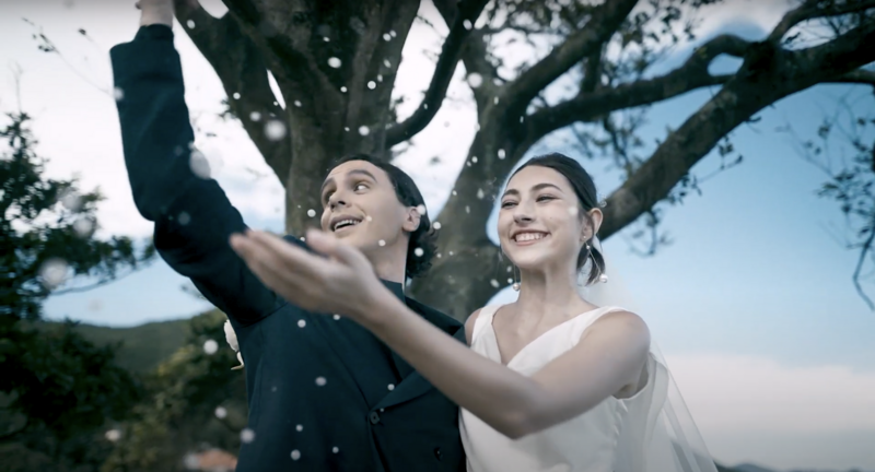 Couple under a large tree reaching upward together, with particles of light floating around them.