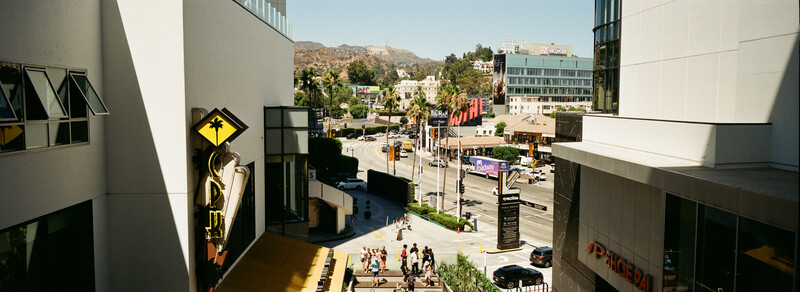 Urban street view from elevated perspective showing downtown plaza with pedestrians, buildings, and mountains in background.