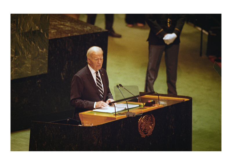 Man in dark suit speaking at wooden podium with microphones in formal assembly hall.