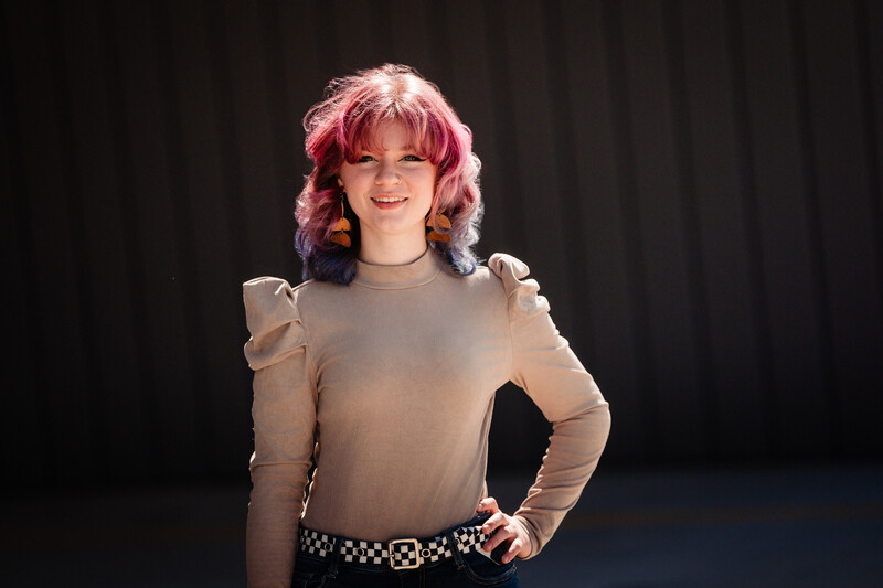 Person with pink wavy hair wearing a beige long-sleeve top and studded belt, posed against a dark curtain background.