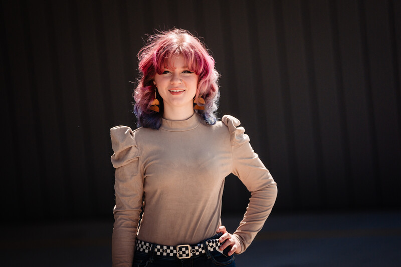 Portrait of a person with pink shoulder-length hair and bangs wearing a beige long-sleeve top with puffed shoulders and studded belt against a dark background.