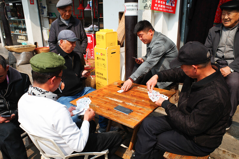 Group of men gathered around a wooden table in a narrow storefront, playing cards or a tile game.