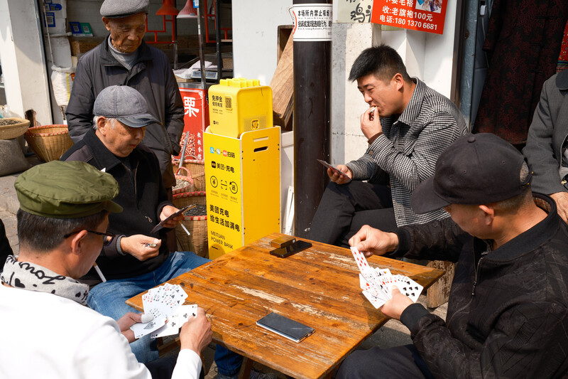 Five men gathered around a wooden table playing cards indoors, with a yellow utility box and storefront visible in the background.