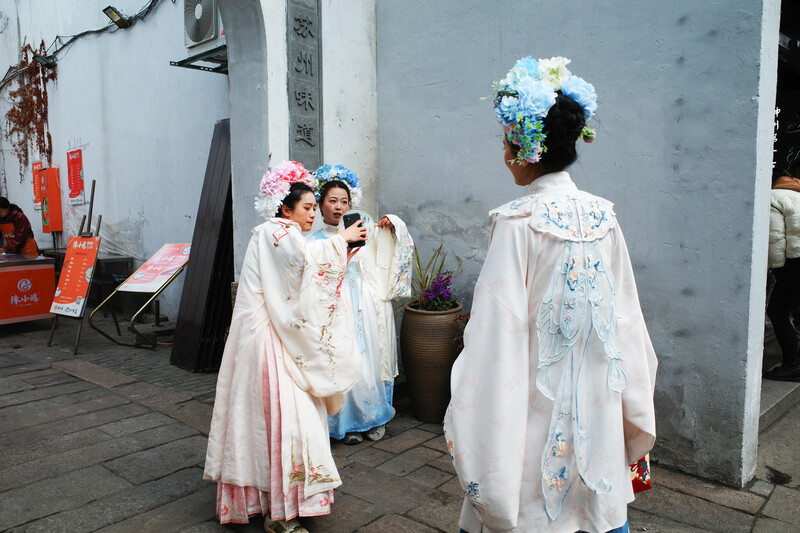 Two people wearing traditional embroidered robes and elaborate flower headdresses stand in a courtyard setting.
