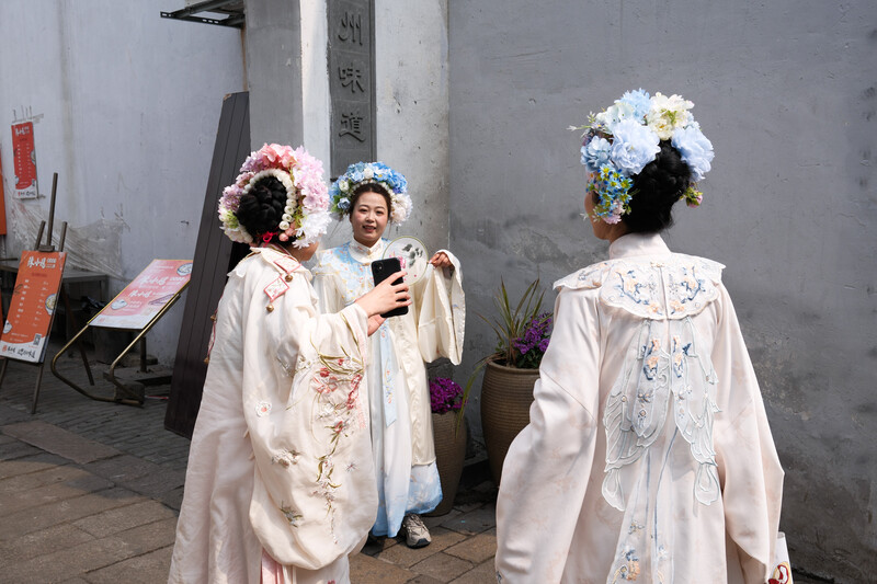 Three people wearing traditional white robes and elaborate floral headdresses stand together indoors.