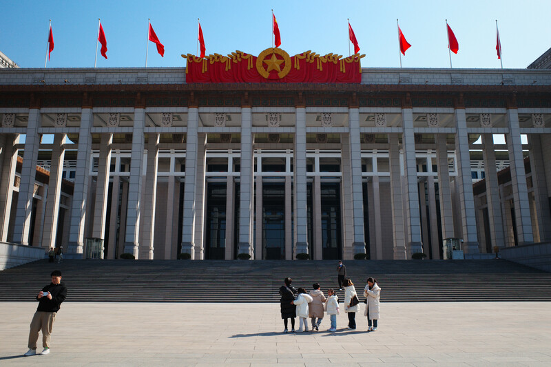 Large government building with white columns and red flags, visitors standing on vast plaza in bright daylight.