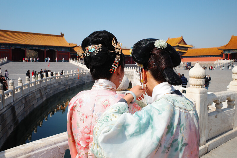 Two women in traditional dress photograph ornate Asian architecture with smartphone.
