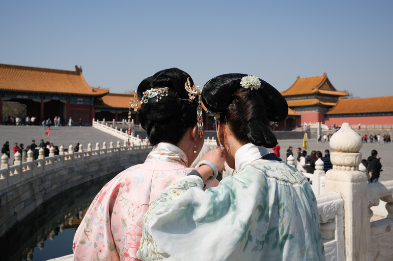 Two women in traditional Chinese dress viewed from behind at a historic palace complex with golden roofs.