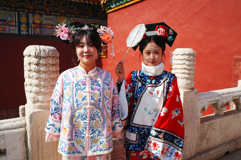 Two young people in traditional Chinese clothing stand before a red wall with decorative stone pillars.
