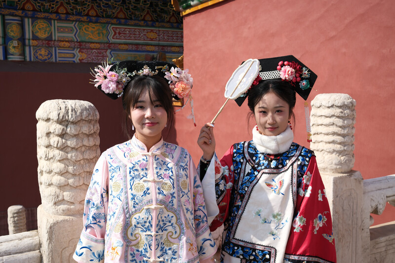 Two young girls in traditional Chinese clothing pose between ornamental stone pillars against a red wall.