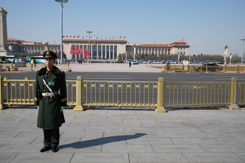 Military or formal uniformed figure standing in a vast plaza with golden barriers and a large governmental building in the background.
