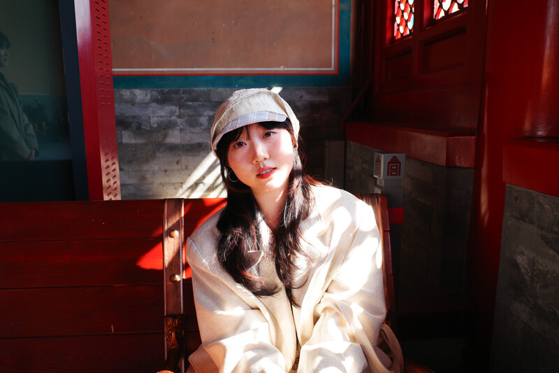 Woman in white jacket and cap sitting against red architectural backdrop with warm directional lighting.