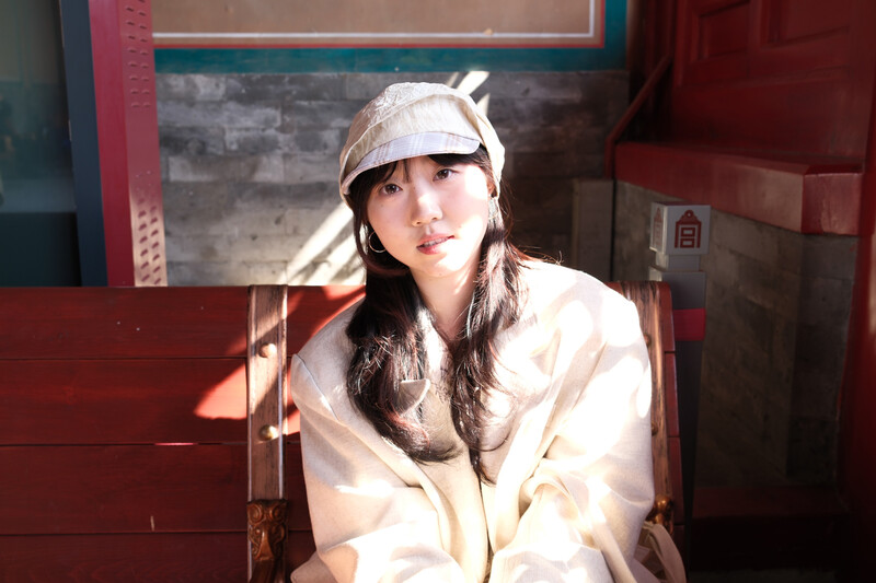 Woman wearing a cream-colored oversized shirt and light cap, seated indoors with warm sunlight creating distinct shadows across her face.