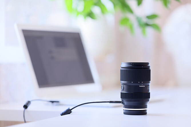 Black camera lens with USB cable connected to white tablet on desk with laptop blurred in background.