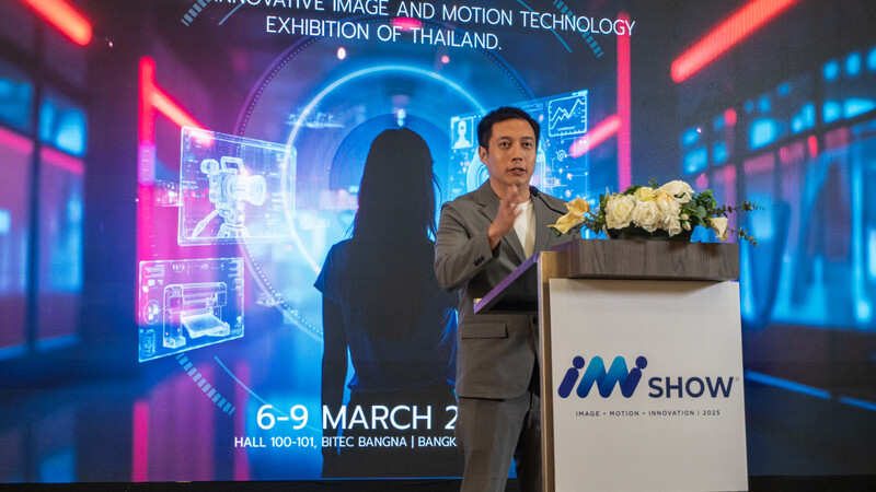 Man in brown blazer speaking at podium with white flowers during tech exhibition with neon blue and pink lighting.