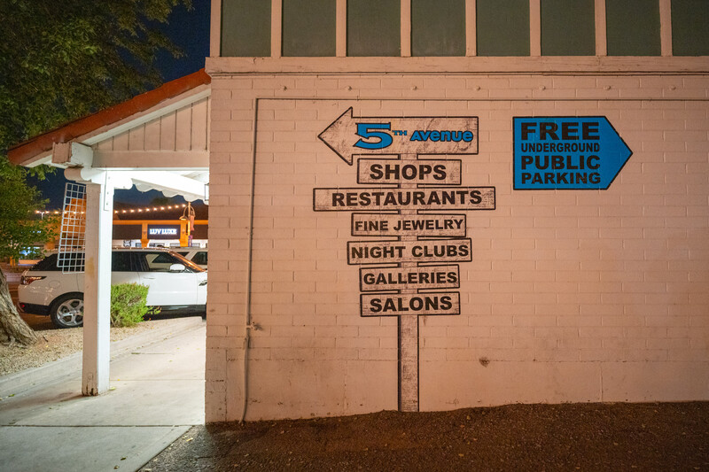 Vintage directional sign painted on brick building wall listing shops, restaurants, and other businesses with arrow pointing left.