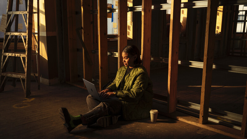 Person sitting on floor working on laptop in dimly lit interior space with wooden beams and windows.