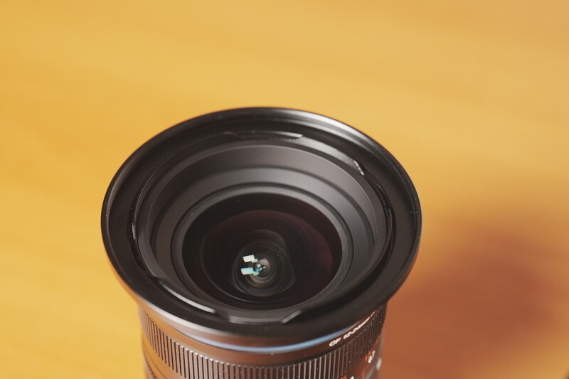 Wide-angle lens with black barrel and glass elements viewed from front against warm yellow background.